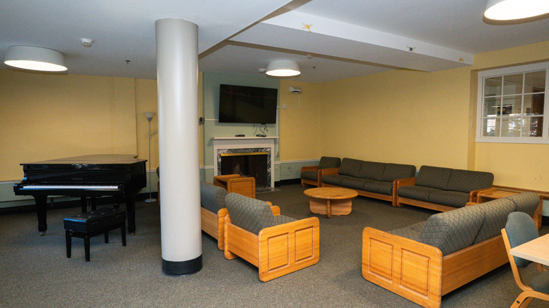 Lounge space with piano in the back left corner and couches surrounding a TV mounted on the wall and fireplace in the back right corner of the room. There are small end tables placed around the couches.