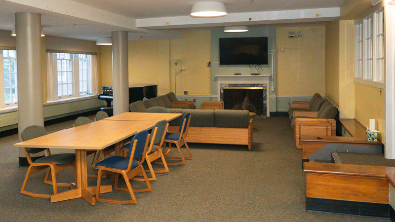 Lounge space with windows along the left wall and couches surrounding a mounted TV and fireplace in the right back corner of the room. There is a piano in the left back corner of the room, and a long table in the middle surrounded by chairs.