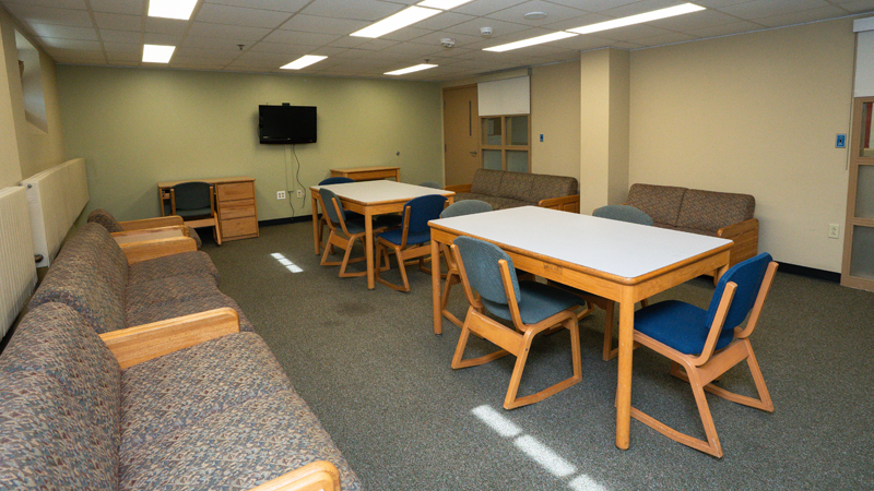 Lounge space with couches along the left and right walls, with rectangle tables surrounded by chairs in the middle of the room. Along the back wall is a table and desk with chair, with a TV mounted on the wall.