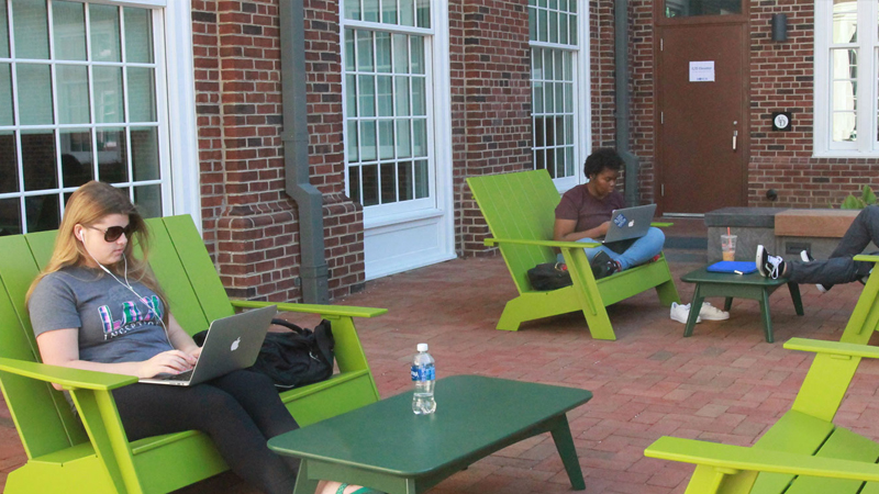 Students sitting at adirondak chairs around tables on their computers and tablets outside the South Academy Residence Hall