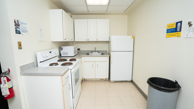 White cabinets with the oven and stove, a microwave on top of the counterspace, sink, and refrigerator. A fire extinguisher is mounted on the left wall, and a trashcan is along the right side of the space.