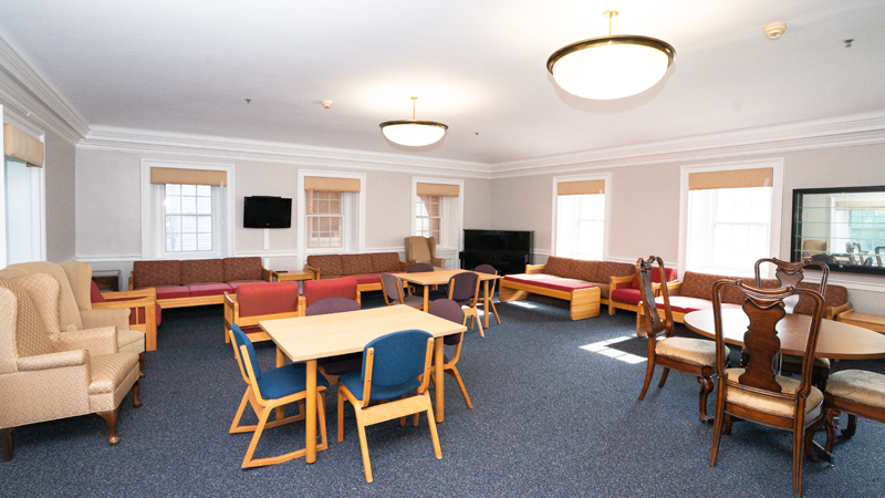 Lounge space with various different tables and chairs around the room. There is a piano in the back corner and a TV mounted on the back wall.