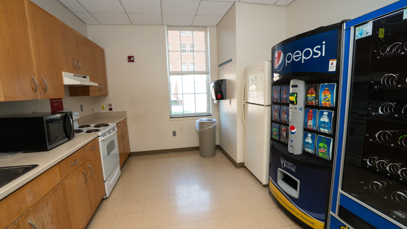 Cabinets along left side wall with sink, microwave and oven and stove. Window on the back wall with a trash can in the corner. On the right side wall is a food and drink vending machine and refrigerator.