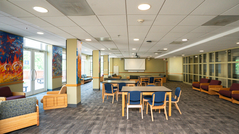 Lounge space with lounge chairs along the left and right walls and various tables and chairs down the midde of the space. There is a projector screen on the back wall, and windows to the inside on the right side and windows visible externally on the left side. 