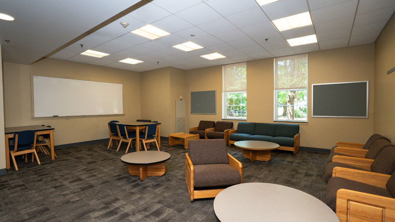 Lounge space with lounge chairs long the right and back walls, and a two tables with chairs along the left wall. There are three small round tables around the middle of the room, and a dry erase board on the left wall, with two push boards on either side of two windows on the back wall.