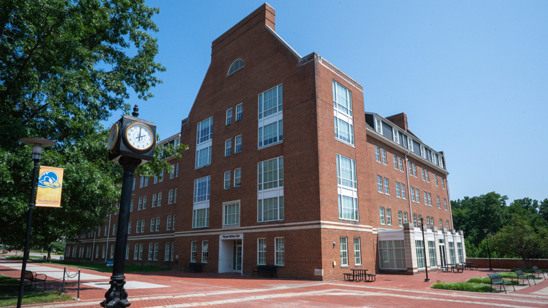 Thomas Mckean Residence hall with a brick walkway, and benches and tables to sit in a courtyard area. A tall clock stands in the middle of the walkway. 