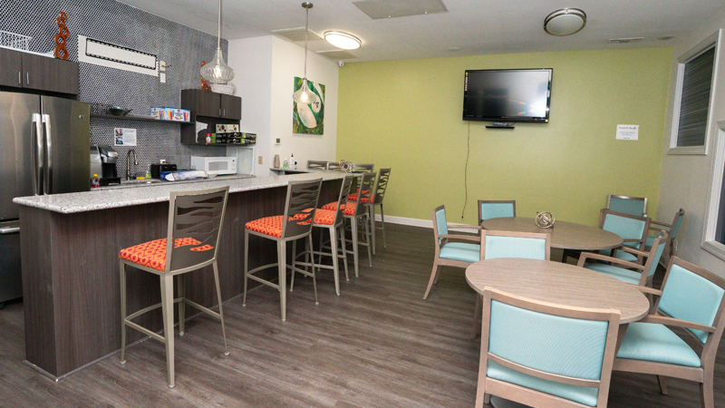 On the left hand side is a refrigerator, sink and then microwave with other miscellanous items on surrounding shelves. In front of the appliances is a counter bar space with eight chairs around it. There is a TV mounted on the back wall, with two chairs in the back corner. Along the right side of the room are two round tables, one with three chairs and the other with four.