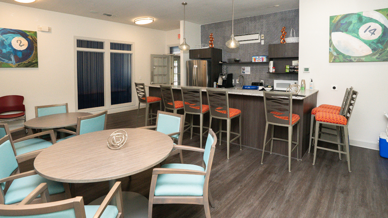 Kitchen space with two round tables - one has three chairs around it while the other has four. Along the back wall are the kitchen appliances, with the top half of the refrigerator visible, a sink and microwave. In front of the appliances is a counter bar space with eight chairs around it. To the left of the fridge is the entry/exit door.