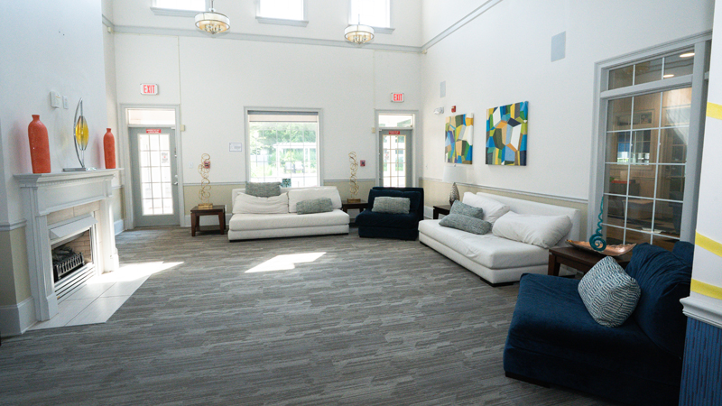 Large common room with high ceilings that has a fire place on the left hand side with decorative pieces on top of the mantel. There is a couch with two pillows along the back wall in front of a large window with two doors on either side. There is a lounge chair with pillow in the corner and then a larger couch with two pillows on the right side wall. In the right corner is another lounge chair with pillow. There are small side tables with decorative pieces on top in between each couch and chair.