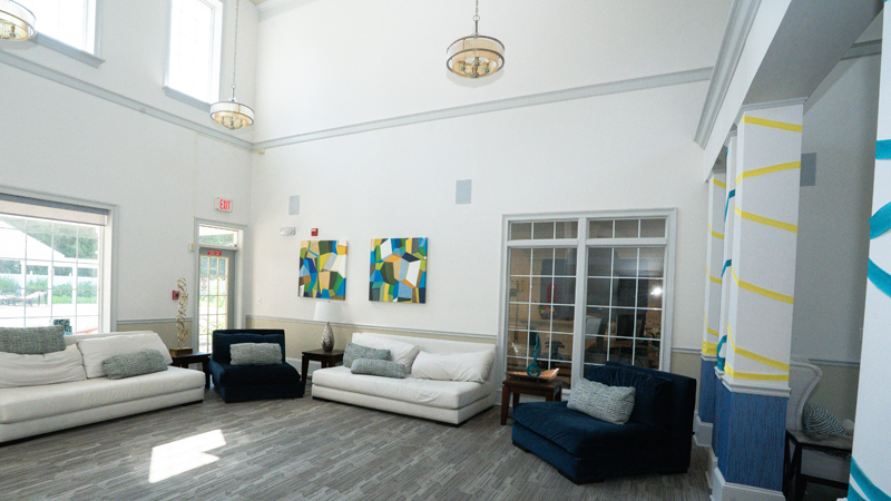 Large common room with high ceilings with couch with two pillows on the left hand side, a lounge chair with one pillow in the corner, and another couch with two pillows along the back wall, with another lounge chair with one pillow in the back right corner. There are small side tables with decorative pieces on top in between each couch and chair. There is a window and door to the outside on the left wall and a tall window into an office along the back wall. 
