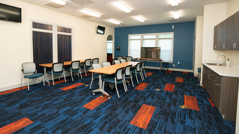 Room with two TVs on the left wall, a TV on a table ahead of three windows on the back wall, and a small counter space with sink, three cabinets above and three cabinets below. There are two tables - one along the right wall with five chairs and the other in the middle of the room with eight chairs around it.