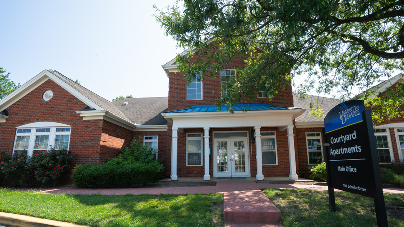 Outside front of the clubhouse space with a set of double doors straight ahead up a brick path with two steps. There is a black sign that says University of Delaware at the top and Courtyard Apartments, Main Office, 100 Scholar Drive on the rest of the sign.