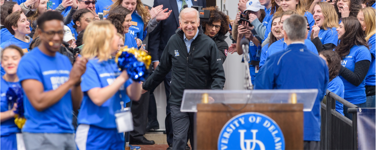 Photo of President Joe Biden walking through the doors of Memorial Hall through a cheering crowd of University of Delaware students, a podium with the UD monogram sits in the foreground.
