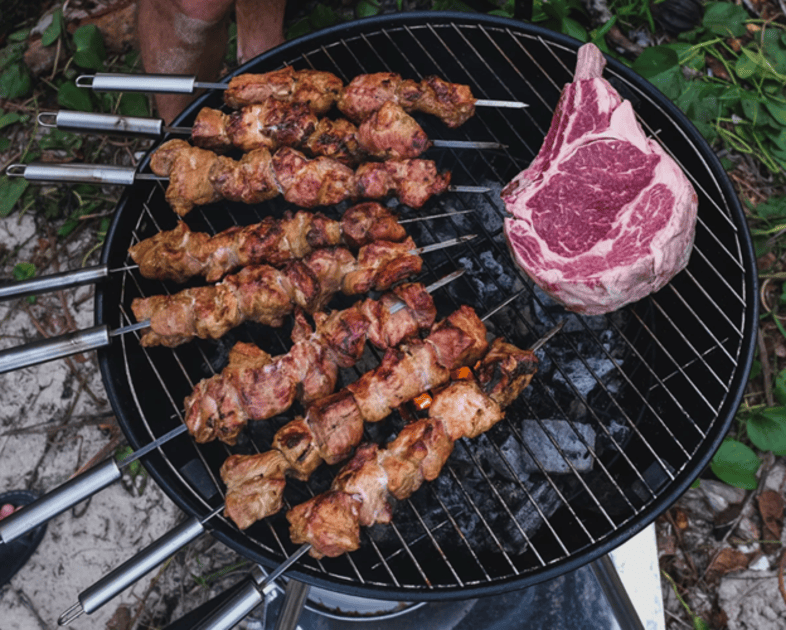 Juicy skewers of marinated meat are grilling alongside a large, raw ribeye steak on a barbecue.
