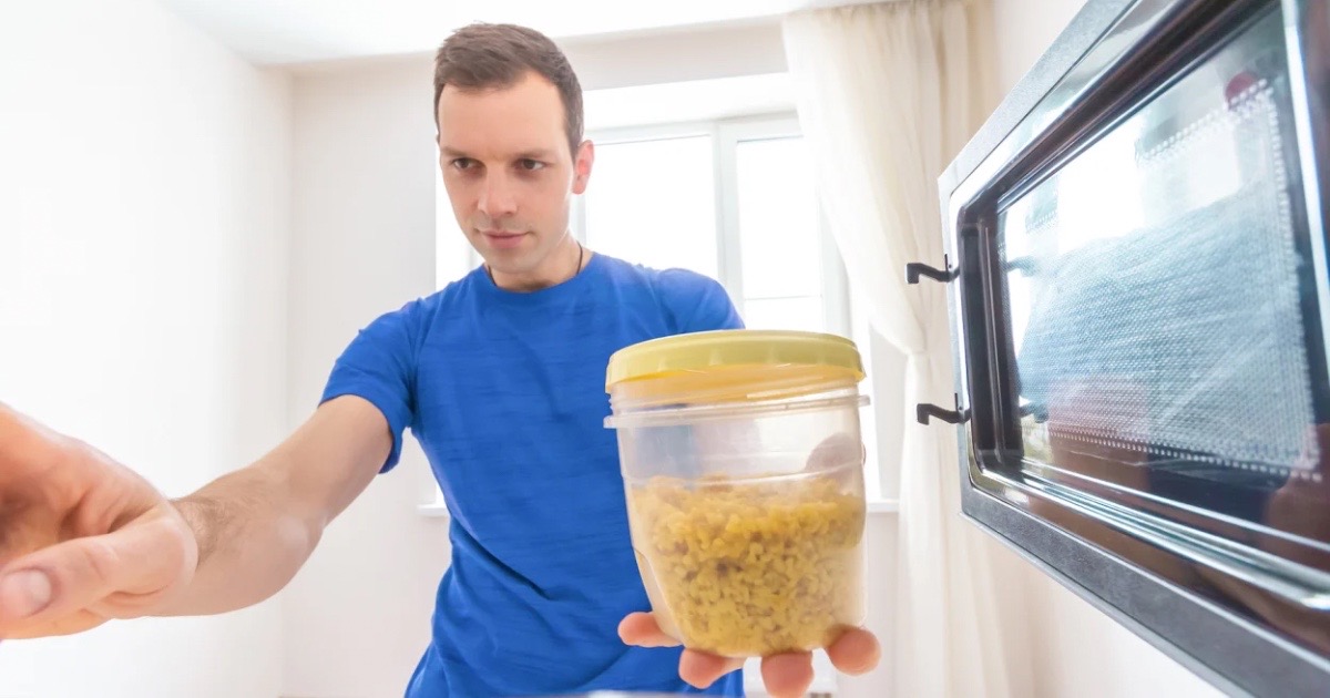 A man in a blue T-shirt reheats rice in the microwave.