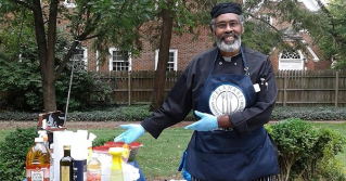 A male Extension volunteer smiling wearing an apron standing next to a table of food.