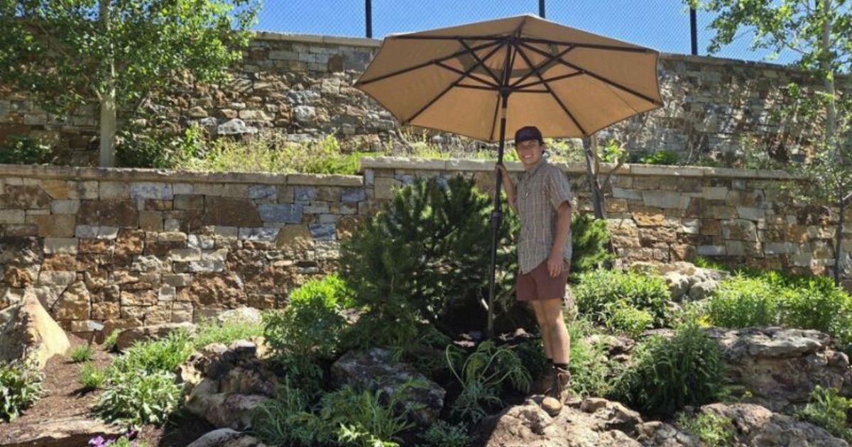 Jackson Fox stands under an umbrella with shrubs next to him, in a garden.
