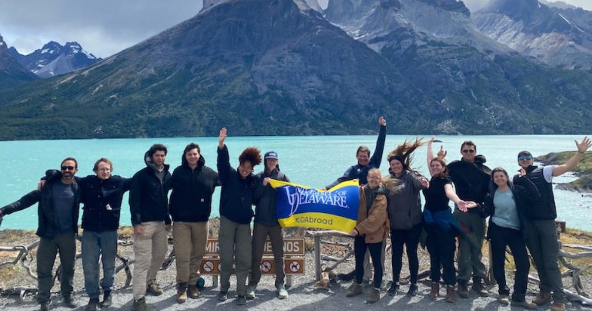 The group of students stands together with a UD flag, a crystal blue lake and mountains in the background.