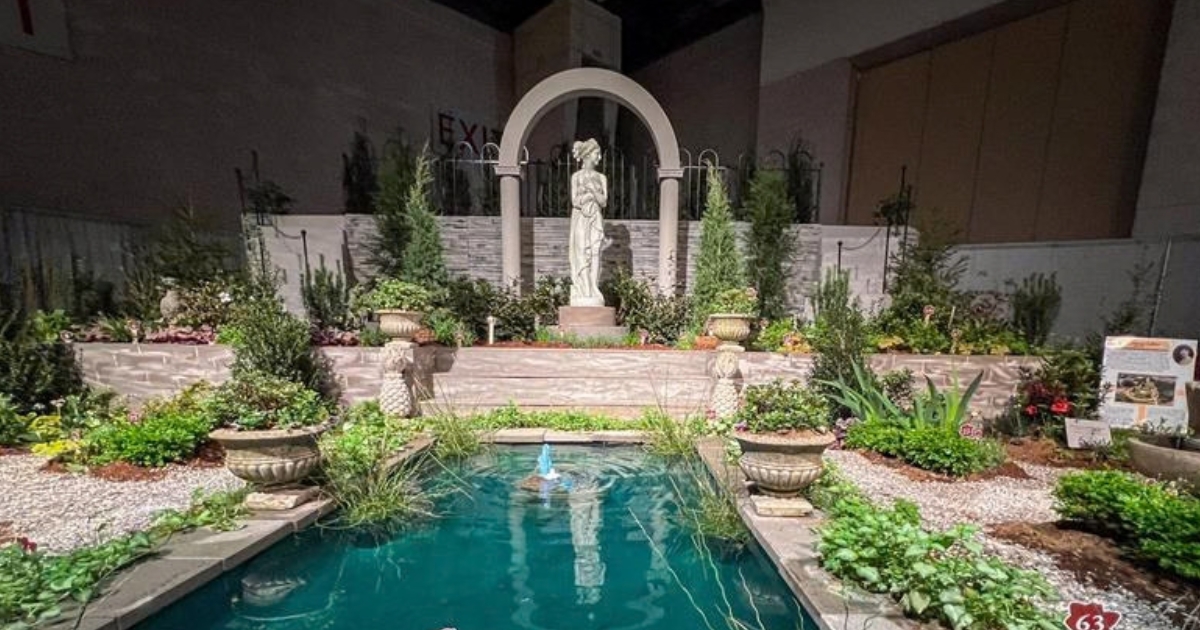 A statue in the back with an arch over it looms over a reflecting pool. Plants are on both sides of the pool.
