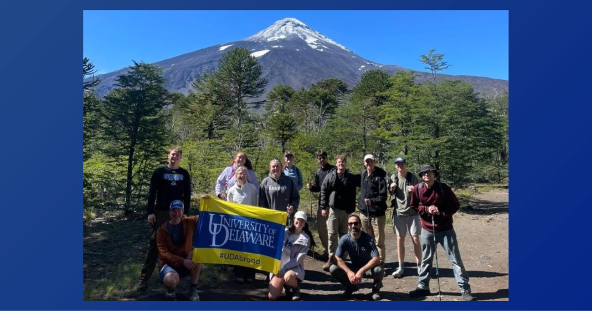 Students hold up a UD flag. A snow-capped mountain sits in the back.