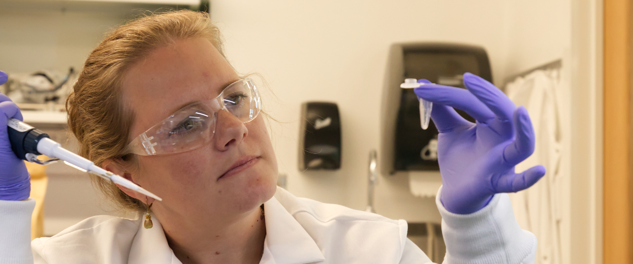 Erin Sparks, wearing a lab coat, gloves, and protective eye gear, examines a sample.