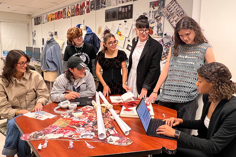 A group of seven students sitting or standing around a red table looking at fashion embellishments and discussing fashion plans