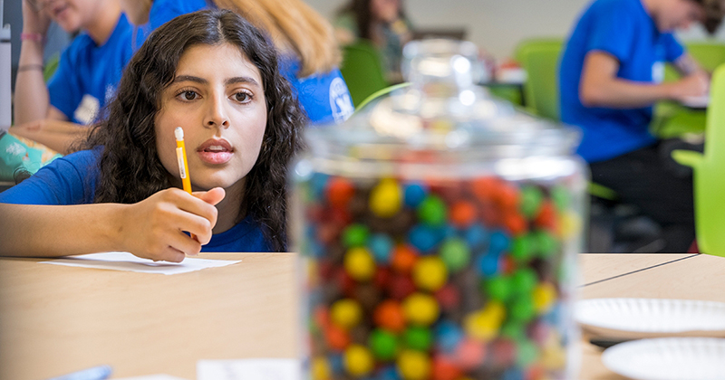 A summer camp participant in a classroom studies a glass jar of colorful candies on a table.