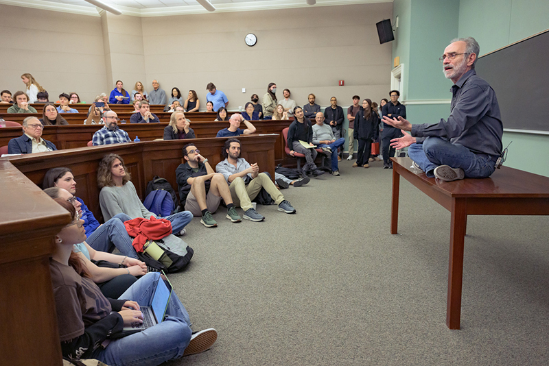 Shelly Kagan, fall 2025 Norton lecture A man sitting on a desk during his speech in front of a crowd of people in a classroom.