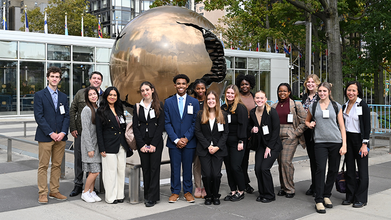 A group of college students in business attire gather in front of a gold globe sculpture in front of the United Nations in New York City.