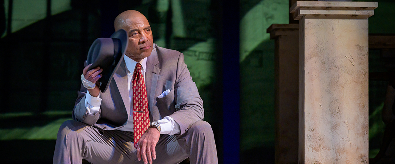 A black man in a suit with a bowler hat sitting on stage