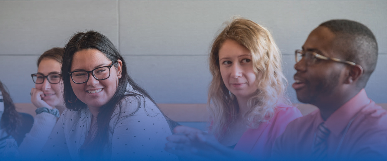 Two college students sit in a classroom and listen attentively to a third student.