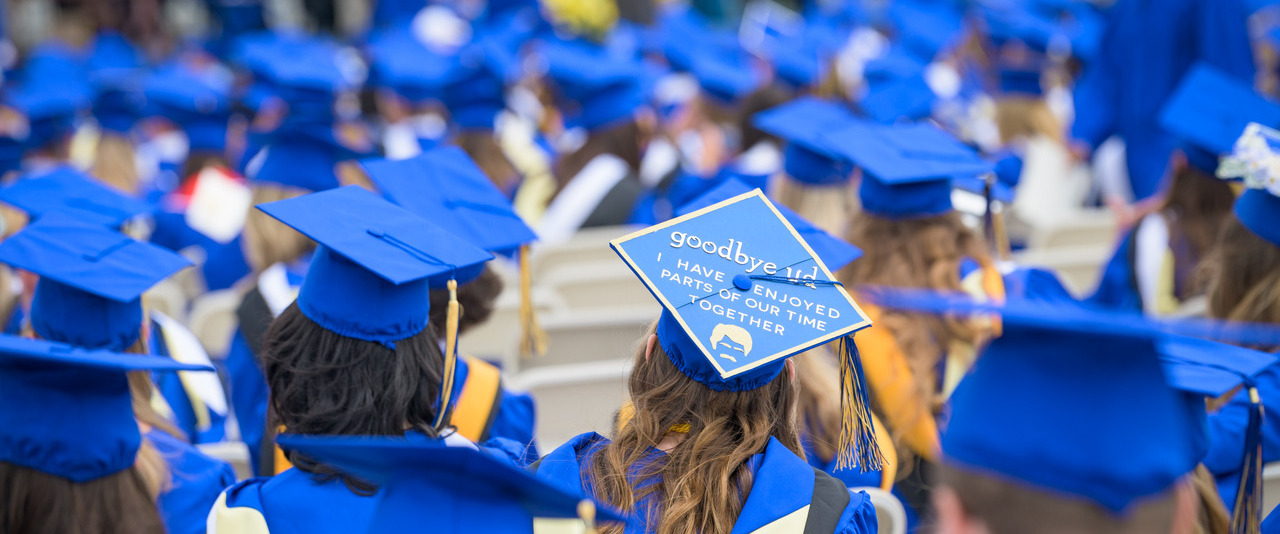Graduates in blue caps and gowns sit at commencement, with one decorated cap reading "goodbye UD, I have enjoyed parts of our time together."