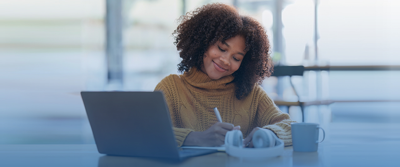 A smiling woman writes in a notebook while sitting at a desk in front of a laptop, headphones and coffee mug.