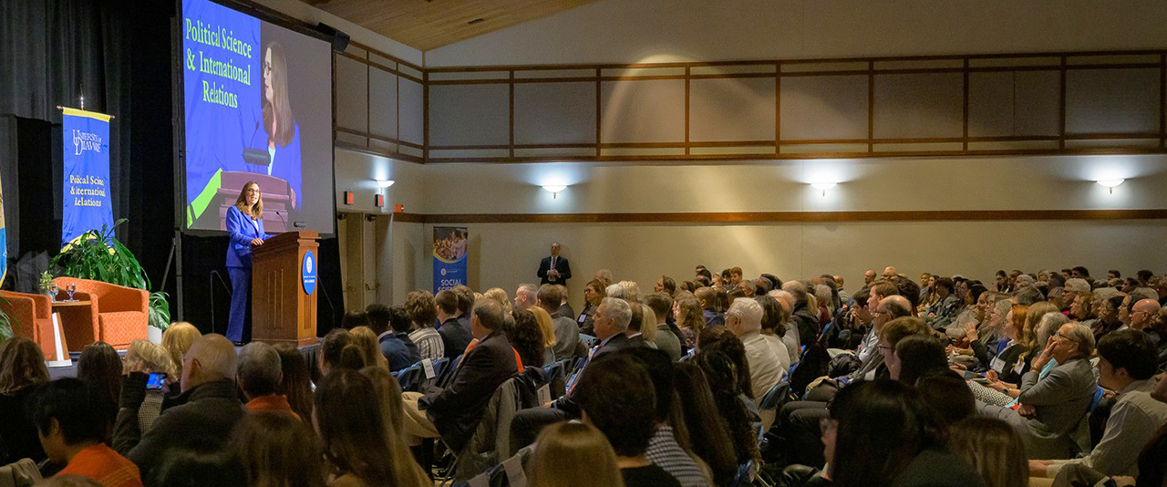 Sarah McBride stands on a stage behind a podium and addresses a large audience at the University of Delaware