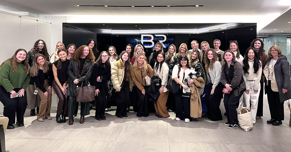 A group of smiling University of Delaware fashion student and faculty gathered for a group photo in the lobby of a fashion company in New York City.