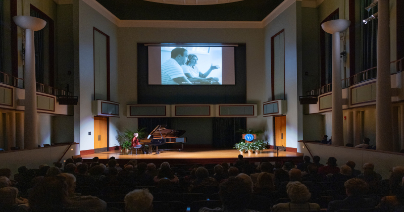 A  musician inside a theater plays a grand piano on a stage in front of an audience.