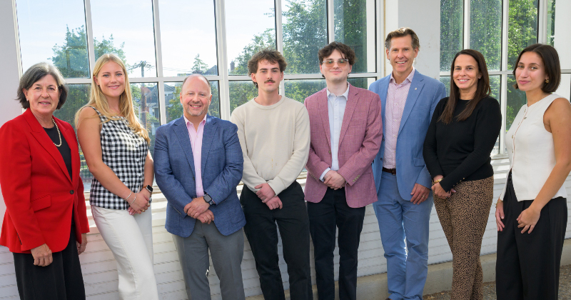 A group of eight smiling people standing in front of a sunny window.