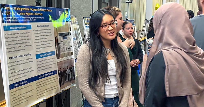 A college student at an event talks to another student while standing next to a poster on an easel.