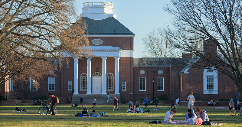 College students gather for picnics and games on a large green lawn in front of a historic brick academic building.