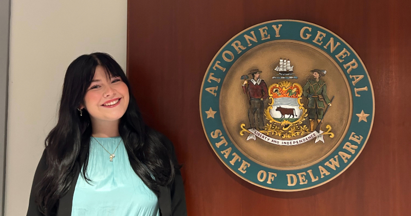 Zoe Quenzel stands next to the seal of the Delaware Attorney General.
