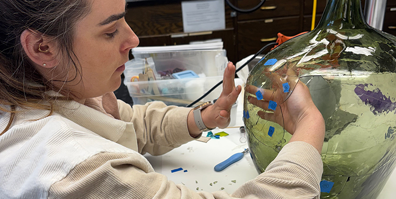 A graduate student in a labaratory reconstructs pieces of a green glass jar with blue tape labeling the pieces. 