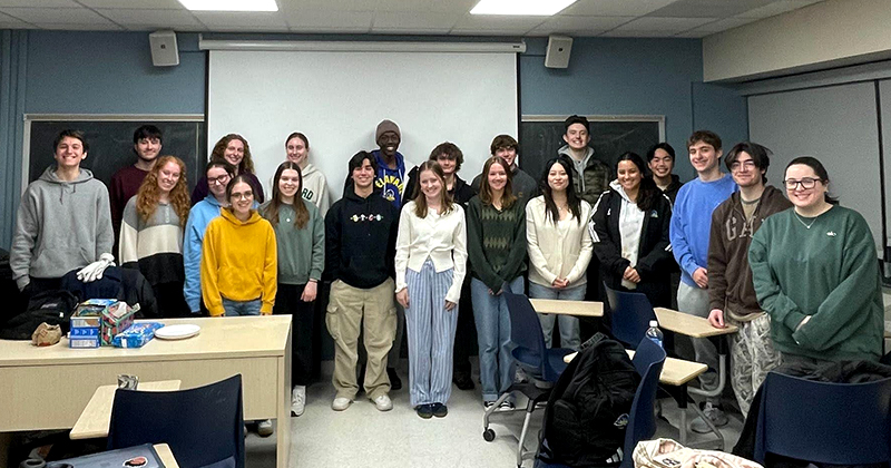 A group of 21 college students stand together inside a classroom for a group photo.