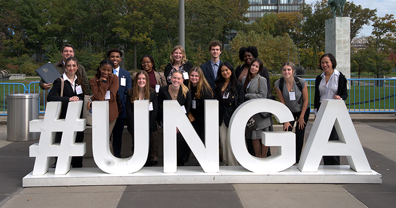 A group of college students and a professor stand behind the #UNGA sign outside the United Nations headquarters in New York City.