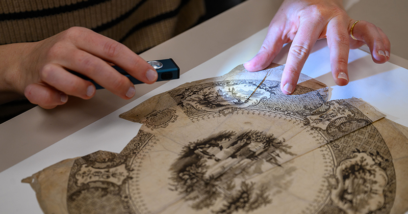 A close-up view of a student's hands holding a light to inspect a 19th century transferware print.