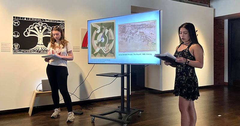 Two college students inside an art gallery deliver a presentation while standing next to a monitor with a slide presentation.