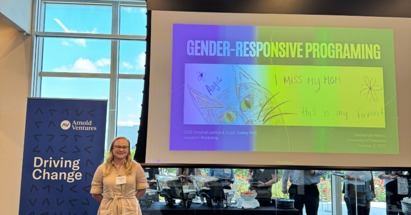 A smiling college student stands next to her slide presentation on a screen behind her.