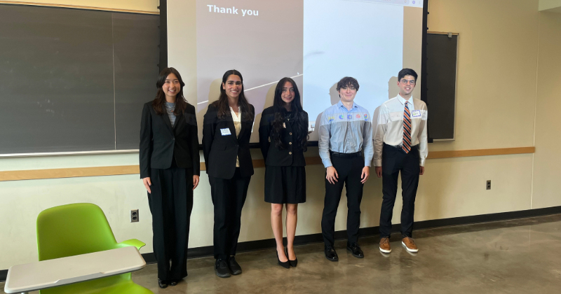 Five college students in business attire stand inside a classroom in front of a projector screen.