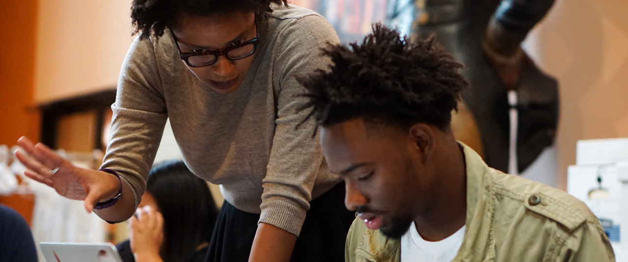 A woman standing and gesturing while helping a man who is seated and focused on a task, possibly on a laptop. 