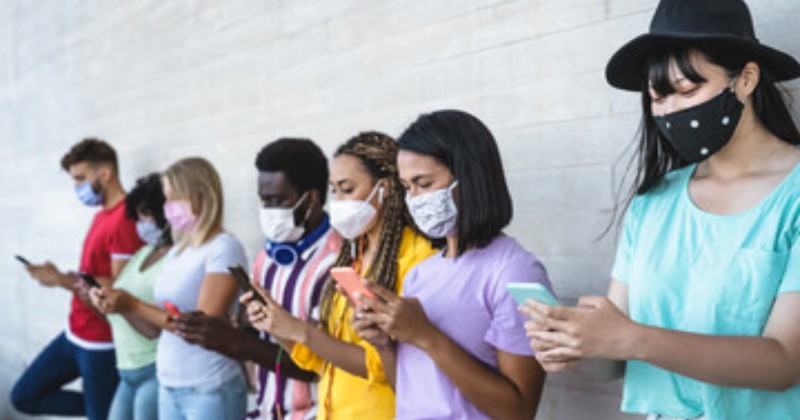A row of seven college students wearing protective face mask and texting on their phones while leaning against a white brick wall