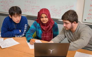 Three college students sit together at a table and look at a laptop computer.